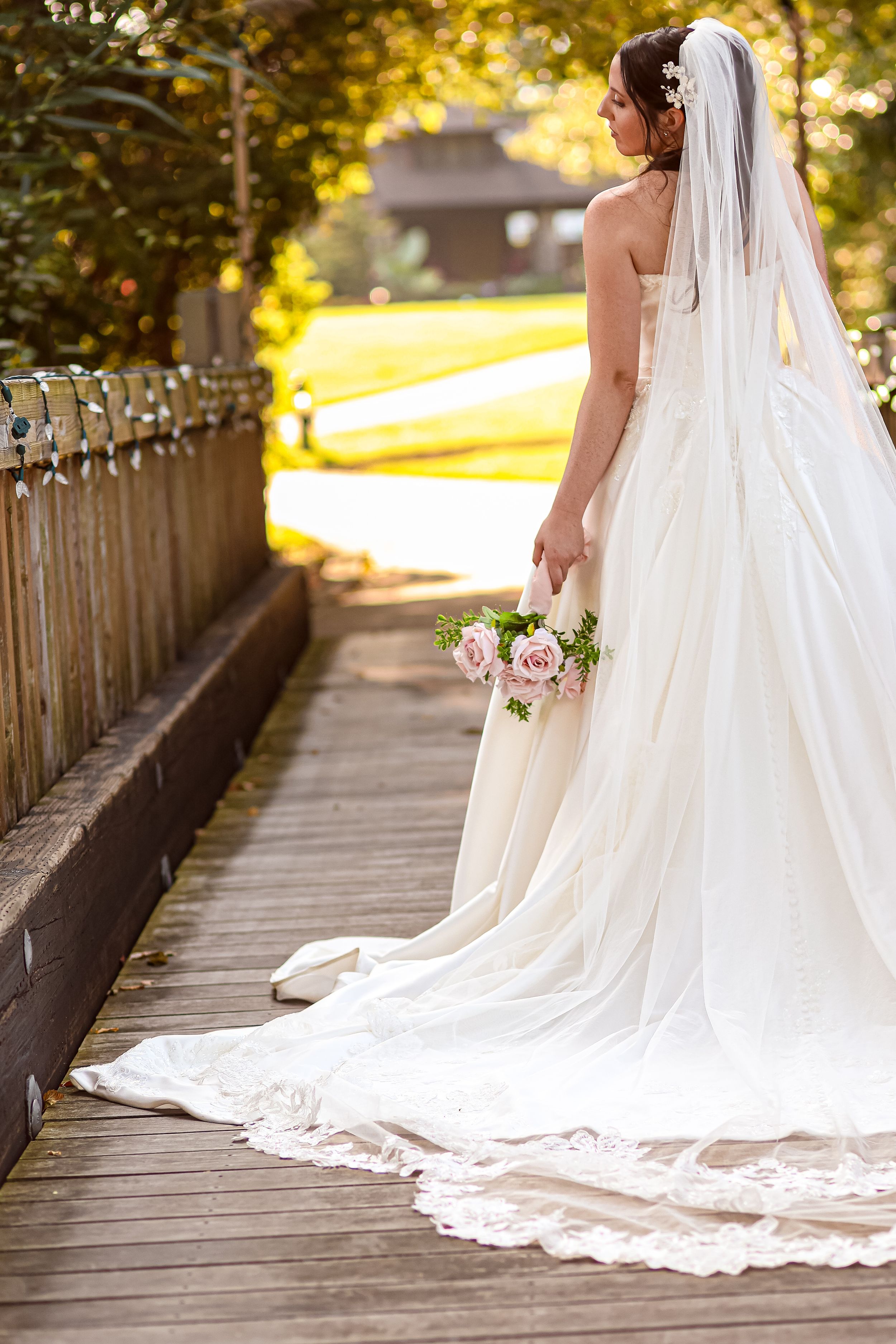 Bride in a strapless white wedding gown with lace-trimmed train and long veil holding a pink bouquet while standing on a wooden bridge.