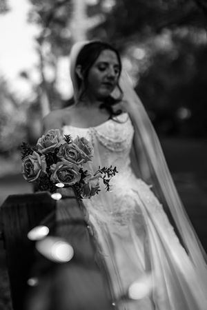 Black and white photo of a bride in a lace wedding gown and long veil holding a rose bouquet outdoors with soft blurred trees.