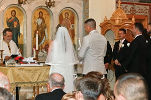 Bride in white gown and veil stands with groom before an Orthodox priest during a wedding ceremony as guests watch inside a church.