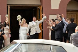Newlywed bride and groom raise their arms in celebration outside a church as cheering wedding guests surround them near a vintage white car.