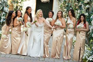 Bride in a white gown laughing with seven bridesmaids in champagne satin dresses holding white bouquets outside a floral-decorated venue.