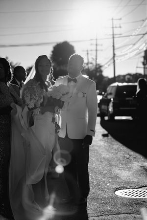Black and white photo of a bride holding a bouquet beside a groom in a white dinner jacket and bow tie, walking down a sunlit street.