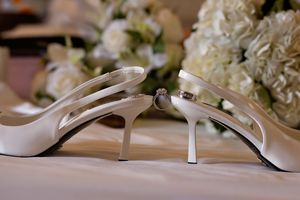 White satin slingback bridal heels with a diamond engagement ring resting on one heel, set before soft white hydrangea flowers.