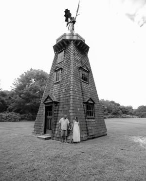 Black and white photo of a couple holding hands at the base of a tall shingled windmill tower with a weathervane on a grassy lawn.