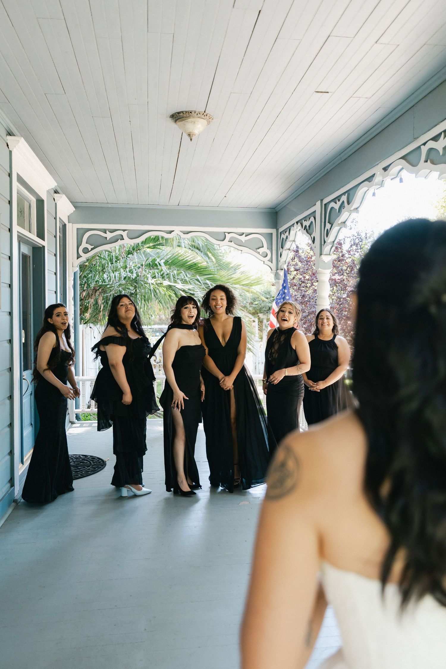 Bridesmaids in black dresses smile on a porch during a first look with a bride in a white gown in the foreground.