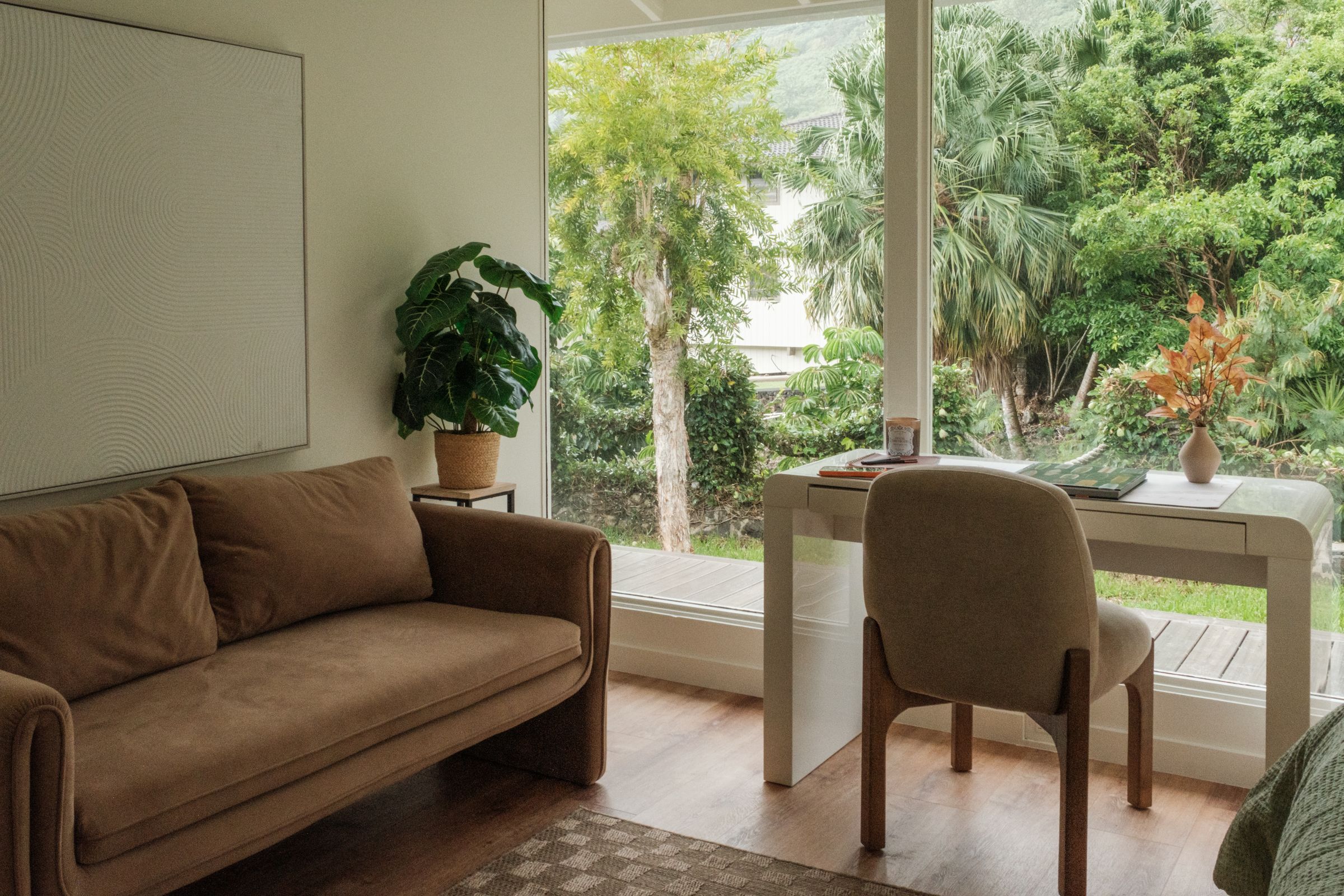 Home office with a brown sofa and a desk facing large windows overlooking tropical greenery.