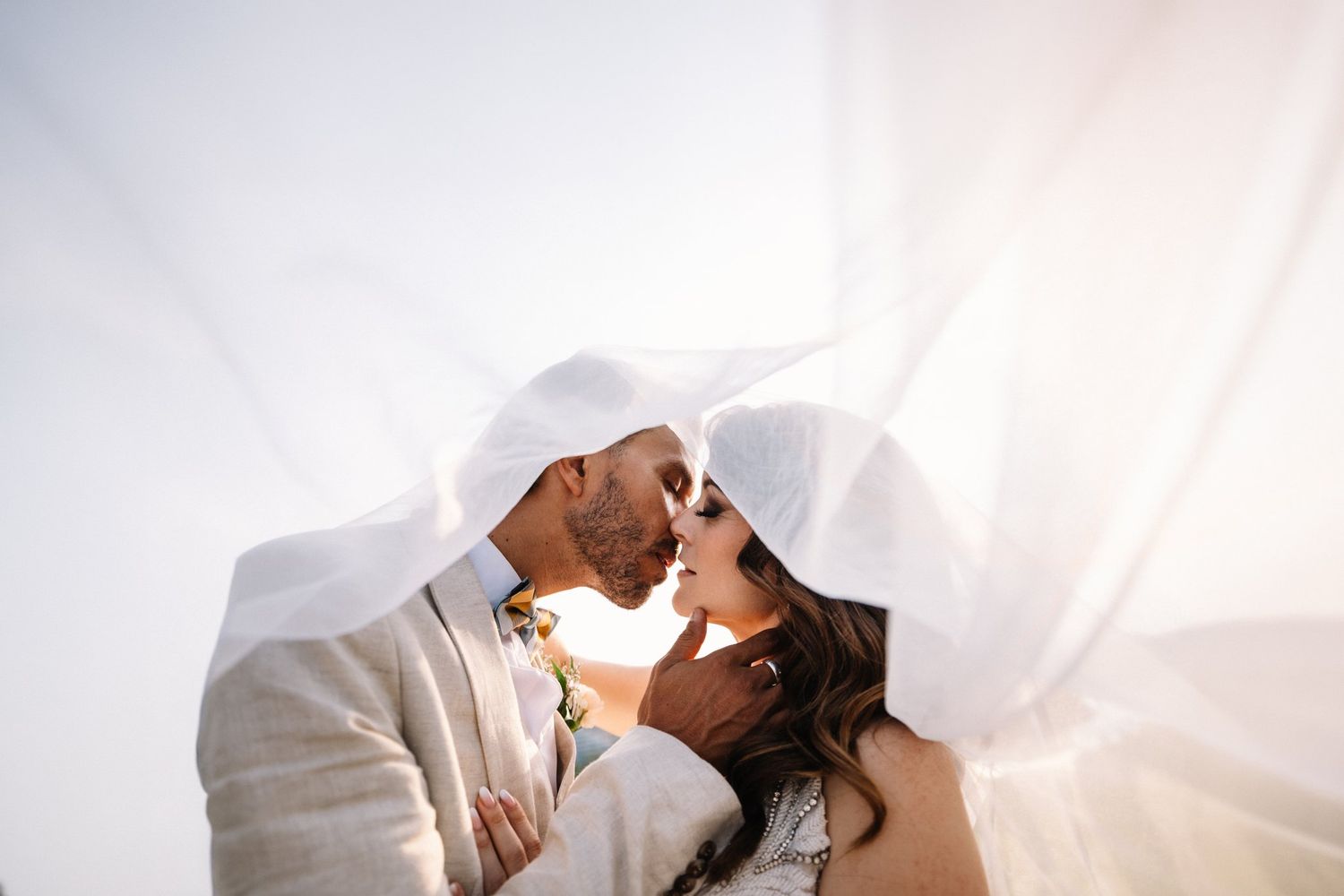 Bride and groom kissing beneath her sheer veil in soft white light