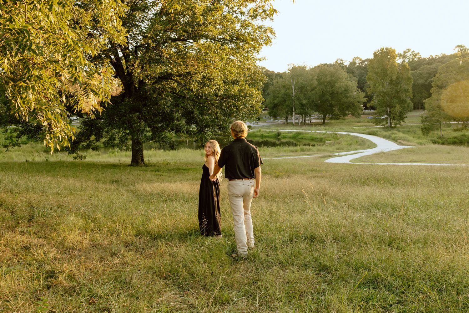 Couple walking hand in hand through a lush green meadow with a winding path and large tree in golden hour light.