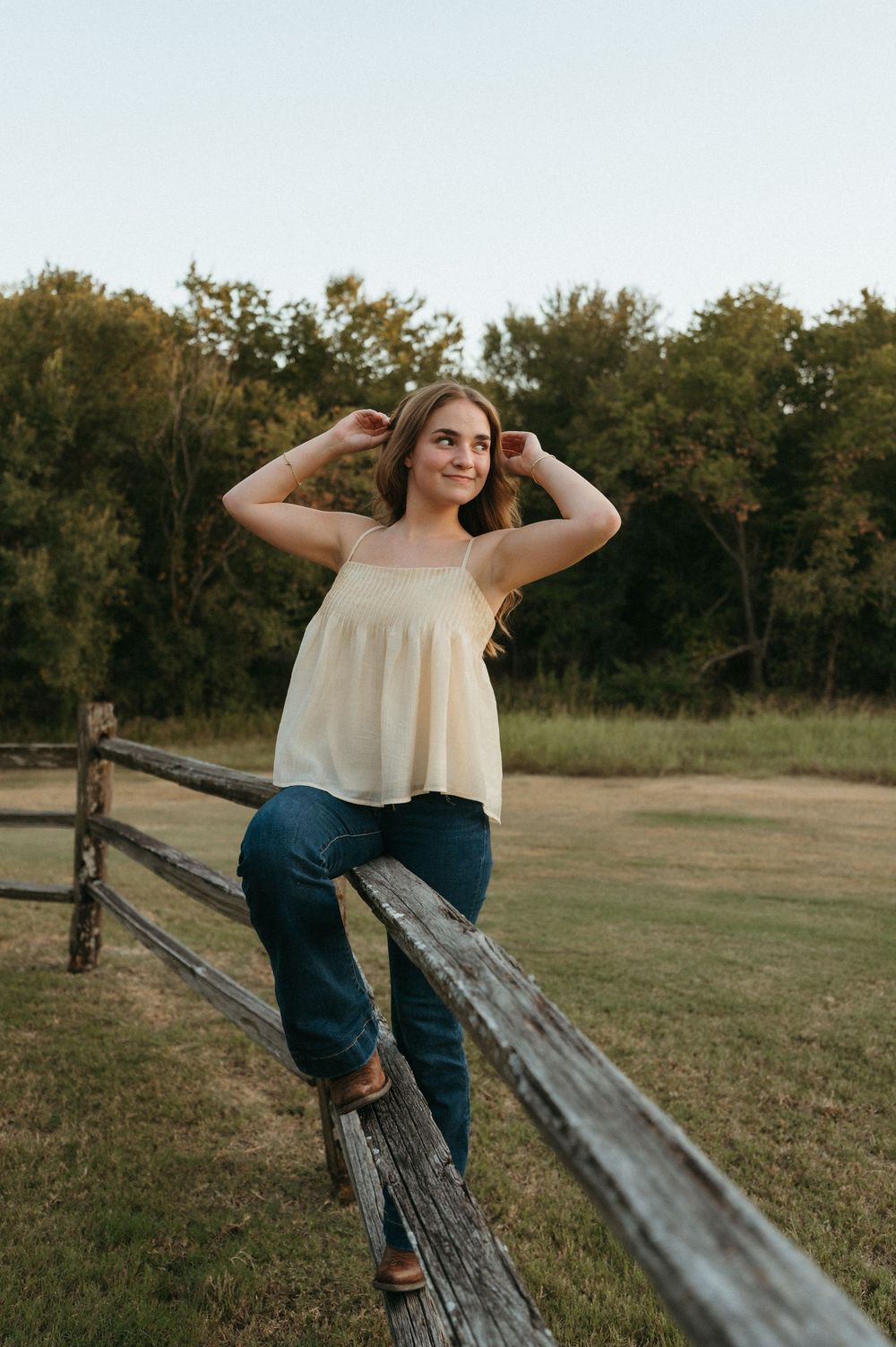 Senior Girl in cream top and jeans leaning on rustic wooden fence in a green field at golden hour.