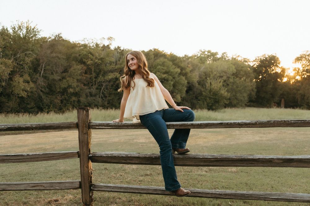 Young woman in jeans and boots poses on rustic wooden fence at golden hour with green trees in background taken by Mary Hickman Photography.