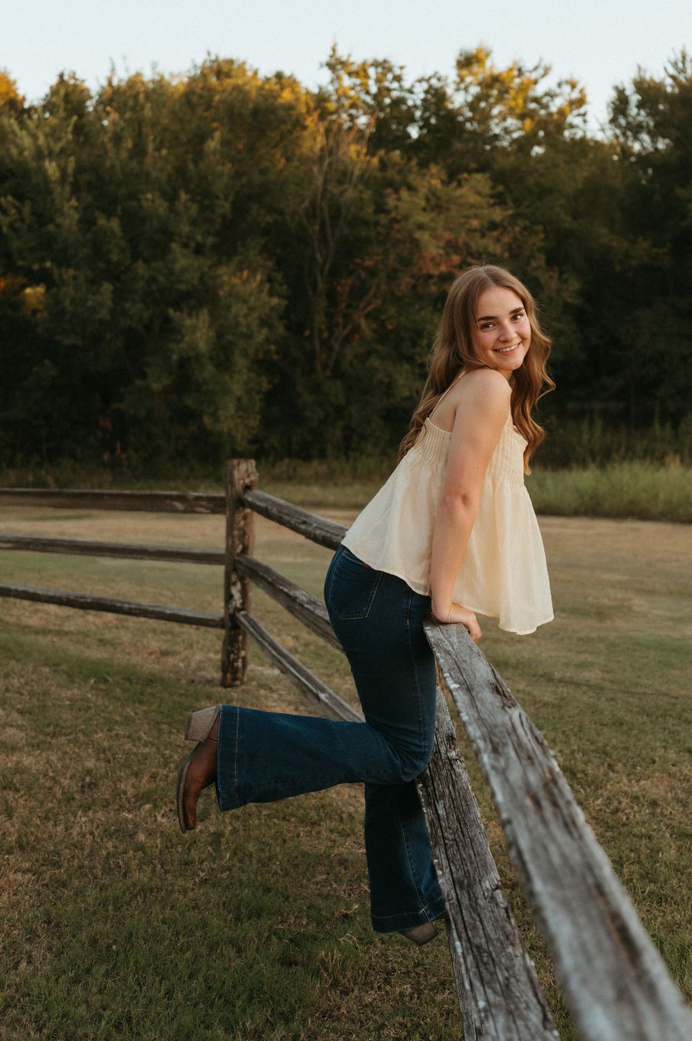 Allen FFA student in white top and jeans leaning on rustic wooden fence in golden hour sunlight with green trees behind her.