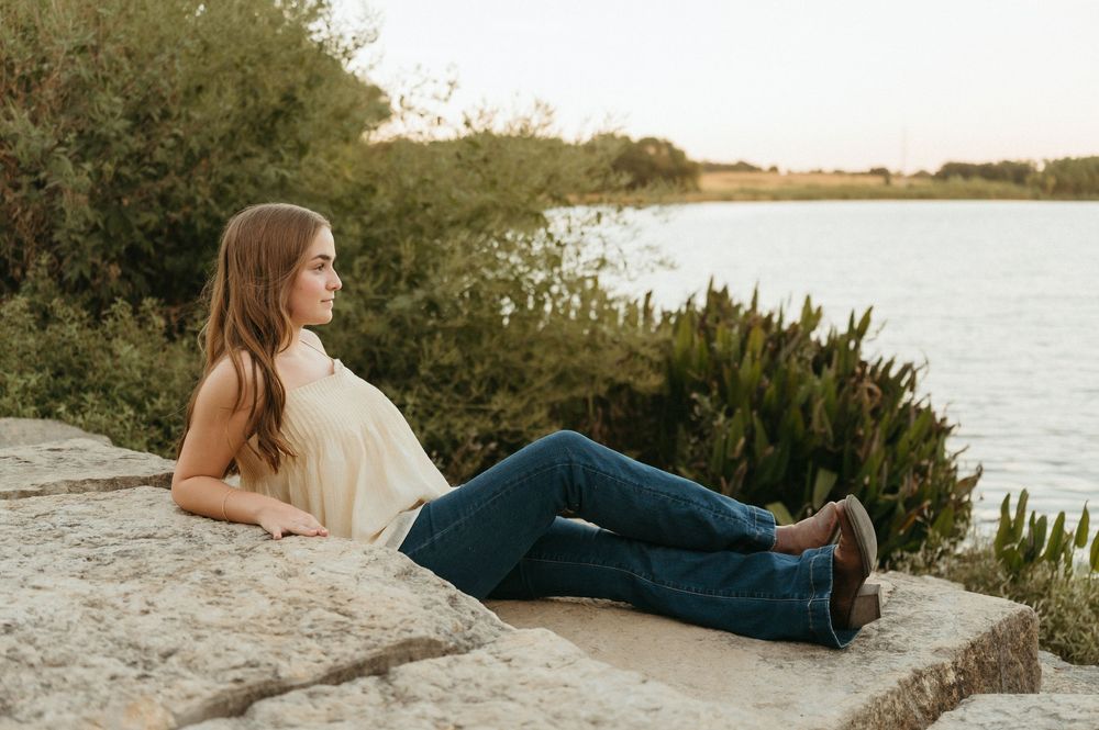 Young woman in jeans and cream top relaxing on stone wall by a lake, gazing into the distance at sunset taken by Mary Hickman Photography