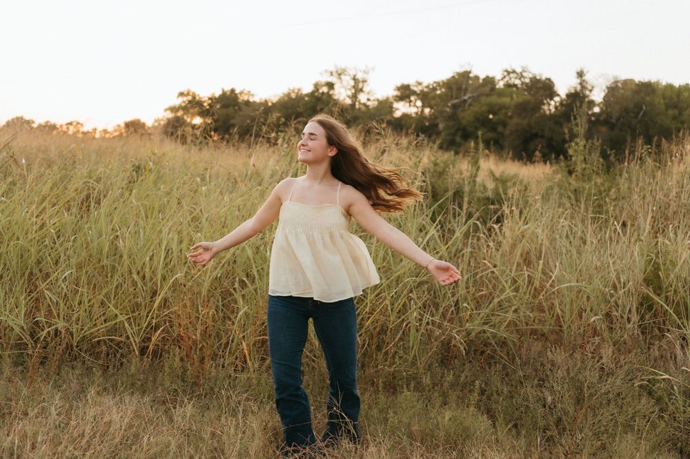 A joyful senior girl with flowing hair stands in a golden field with arms outstretched at sunset in Plano, TX