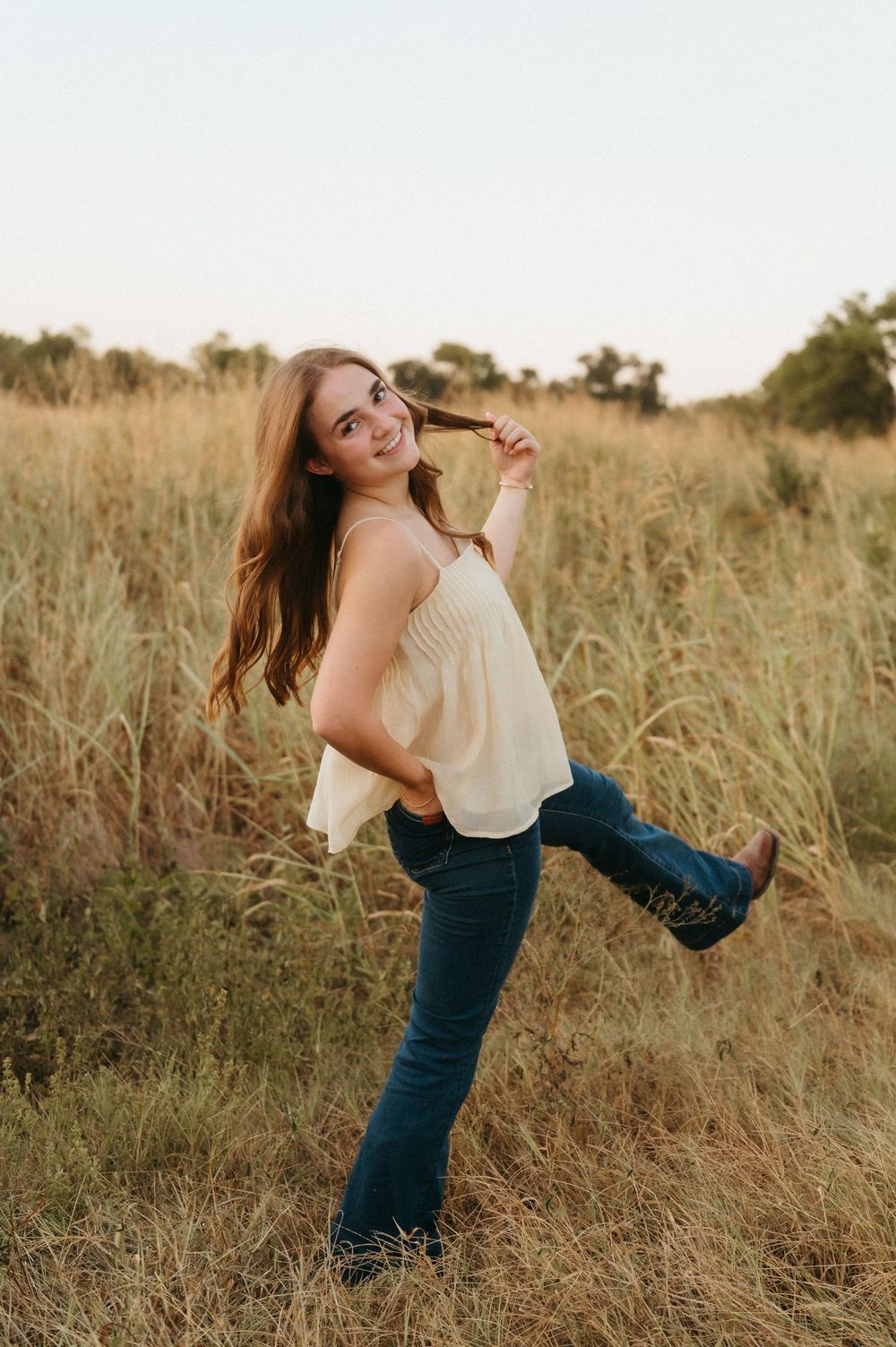 Young woman in white top, jeans, and cowboy boots posing playfully in a golden grass field at sunset at Oak Point Plano.
