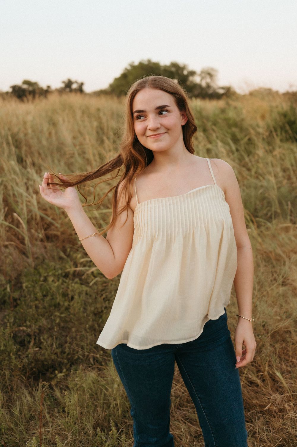 Senior girl stands smiling in golden field, twirling her long brown hair taken by Mary Hickman Photography.