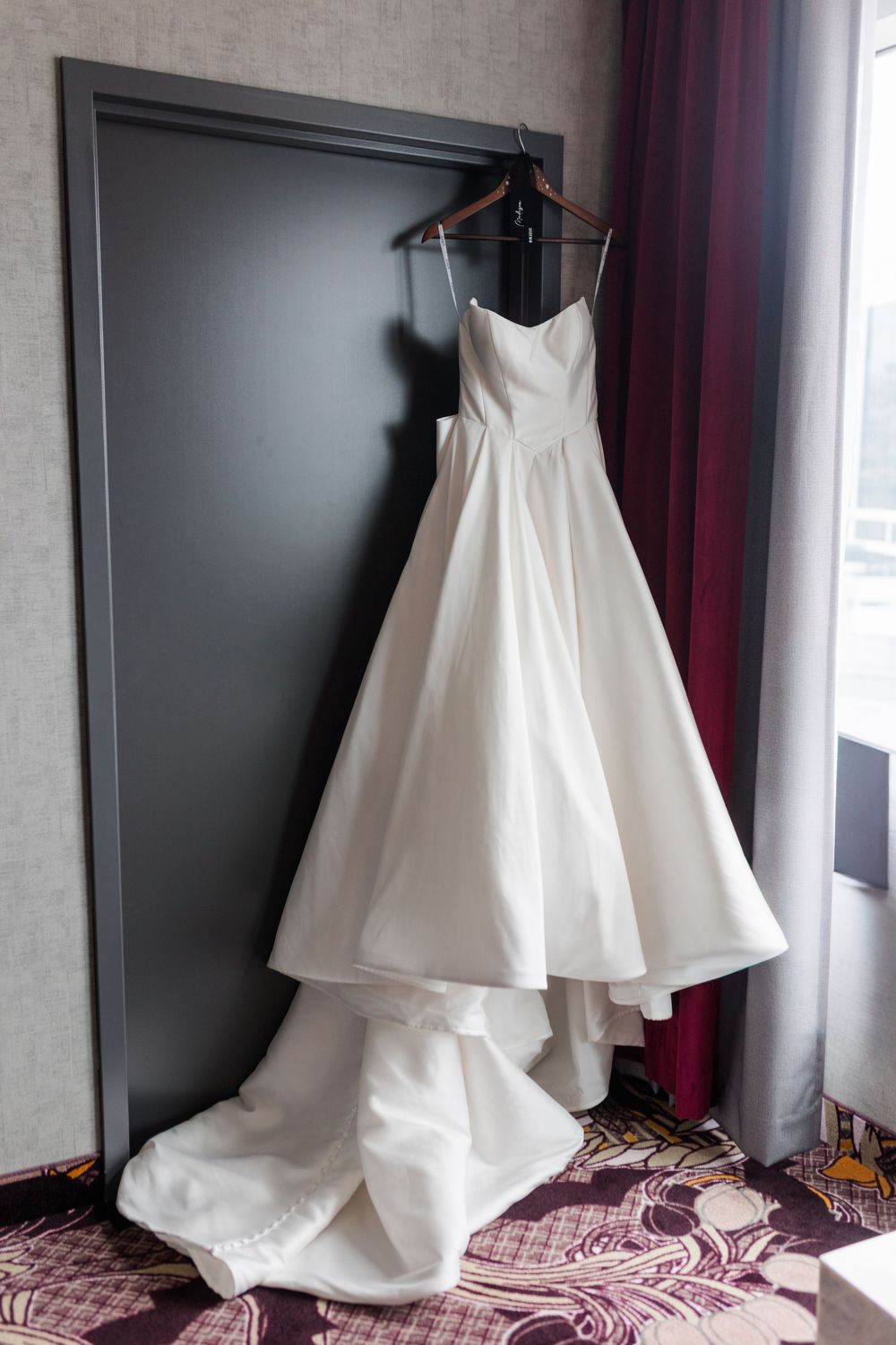 Elegant white strapless A-line wedding dress with a long train hanging on a wooden hanger against a gray door.