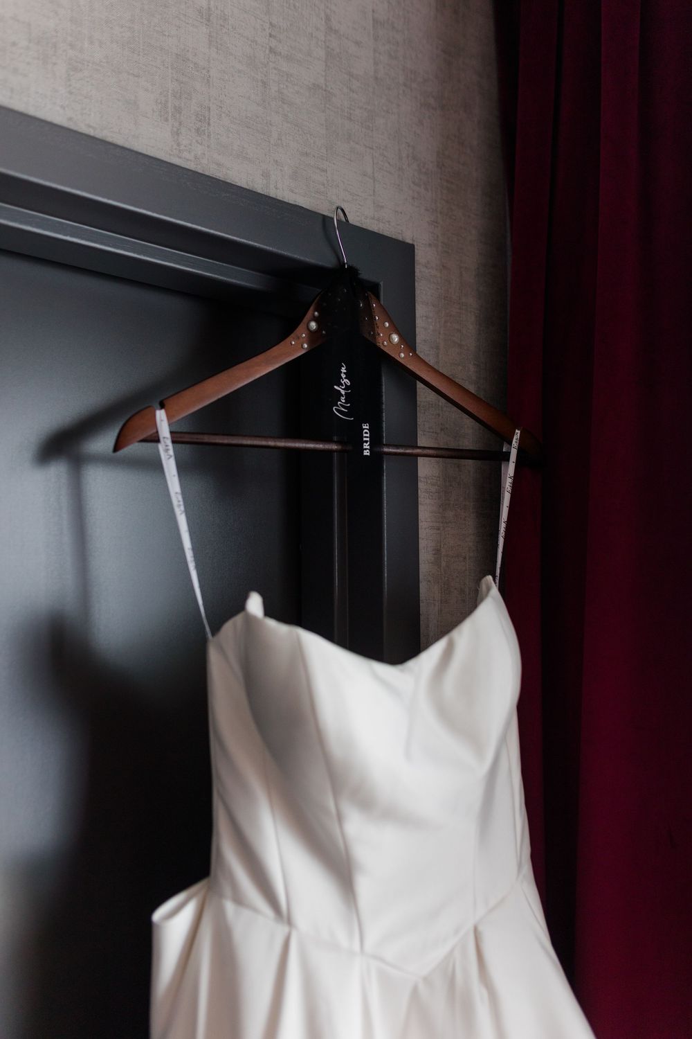 White satin wedding dress hanging on a personalized wooden hanger against a dark gray wall with red curtain.