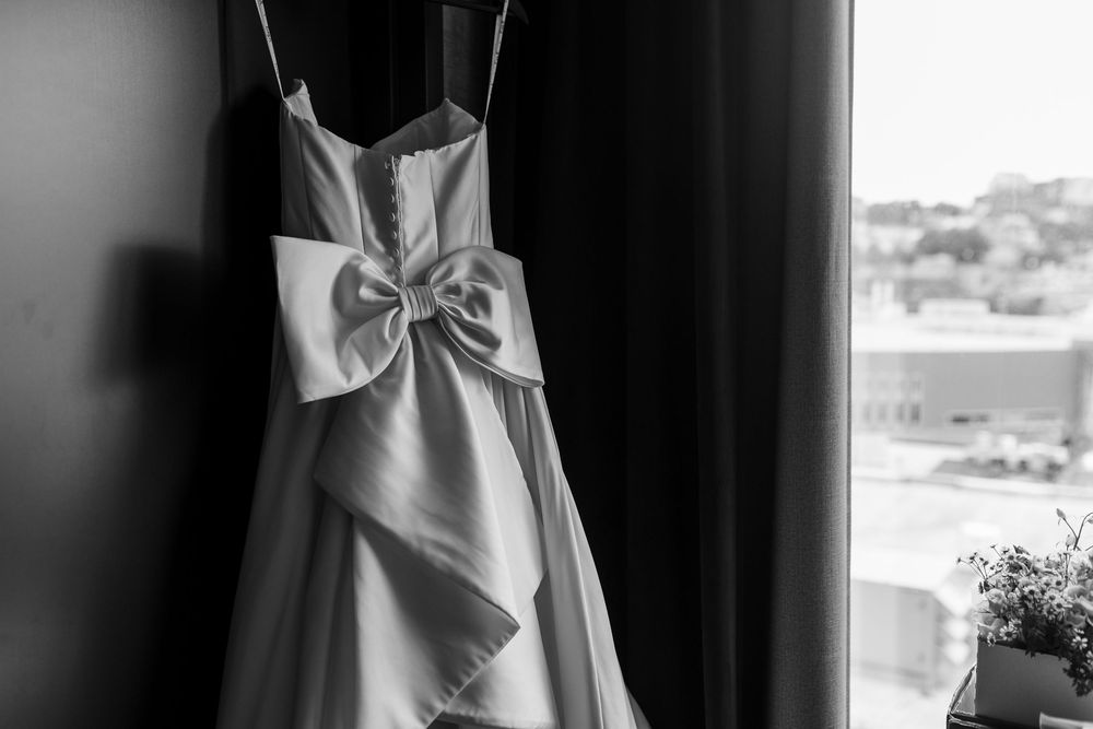 Black and white photo of an elegant strapless wedding dress with a large bow, hanging near a window.