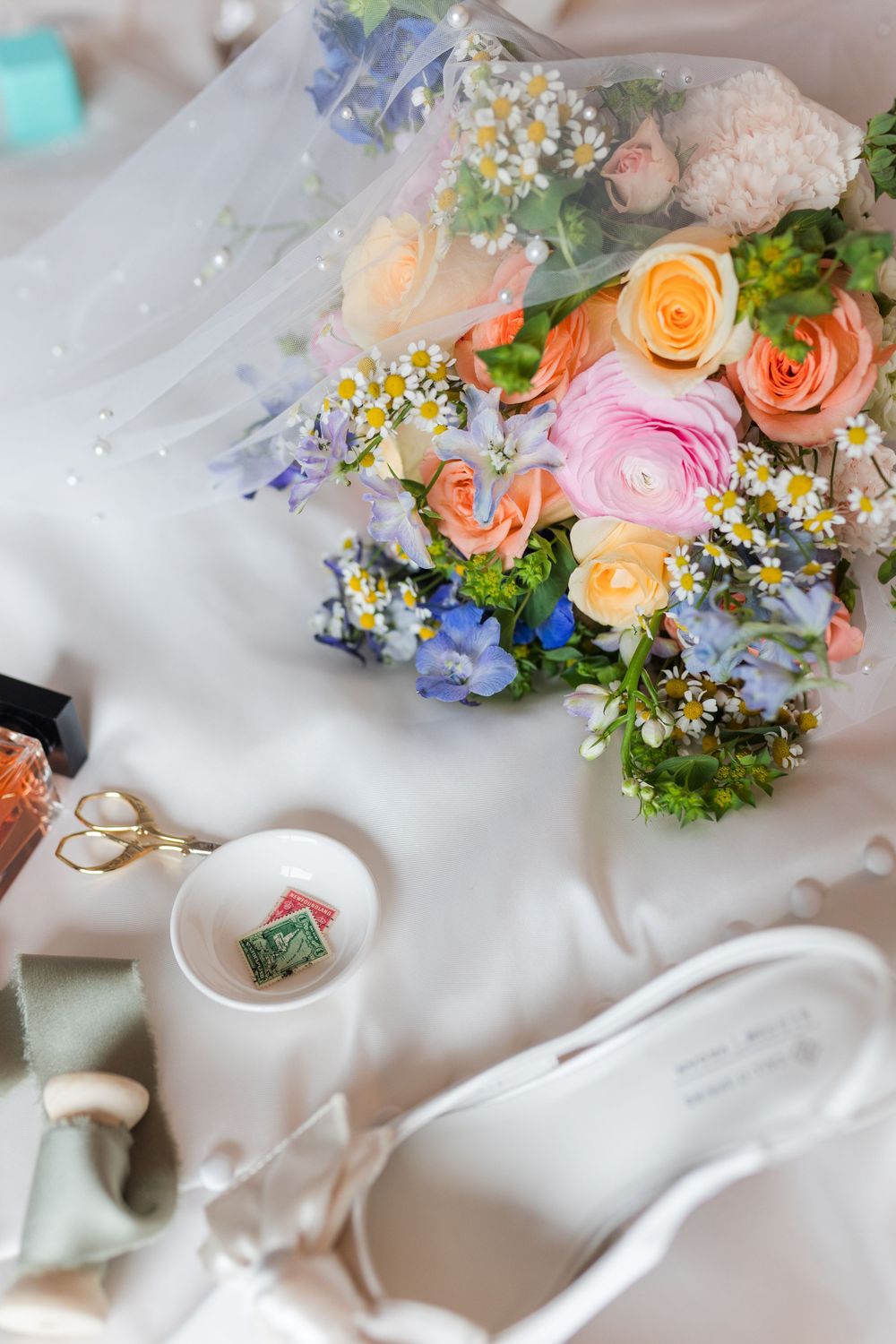 Bridal flat lay with colorful bouquet of roses, white heels, gold scissors, and ring dish on white fabric.