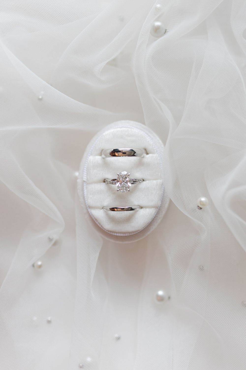 Three wedding rings displayed in a white velvet ring box on a pearl-adorned bridal veil.