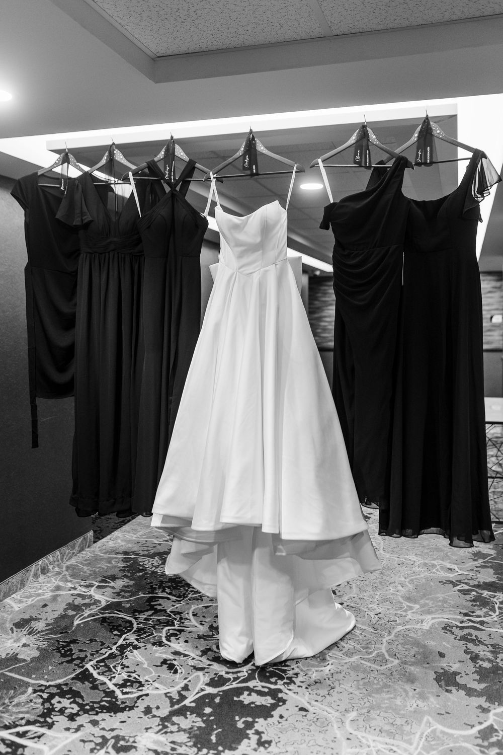 Black and white photo of a white wedding gown flanked by dark bridesmaid dresses hanging on a rack.