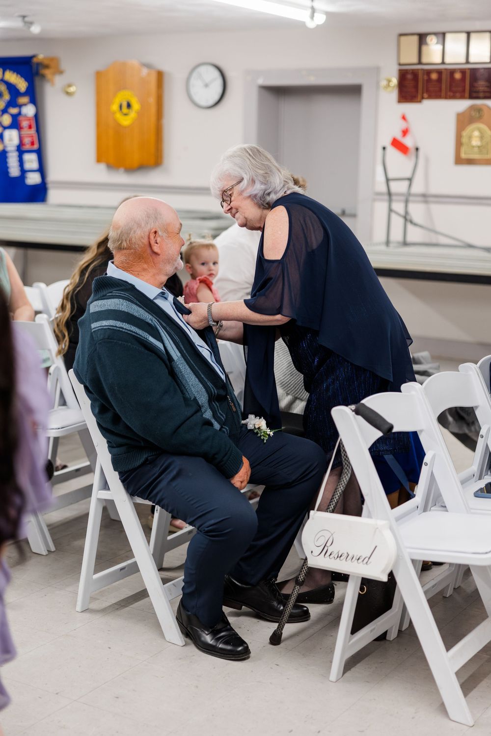 Elderly couple sharing a tender moment at a wedding reception, seated on white chairs in a decorated hall.