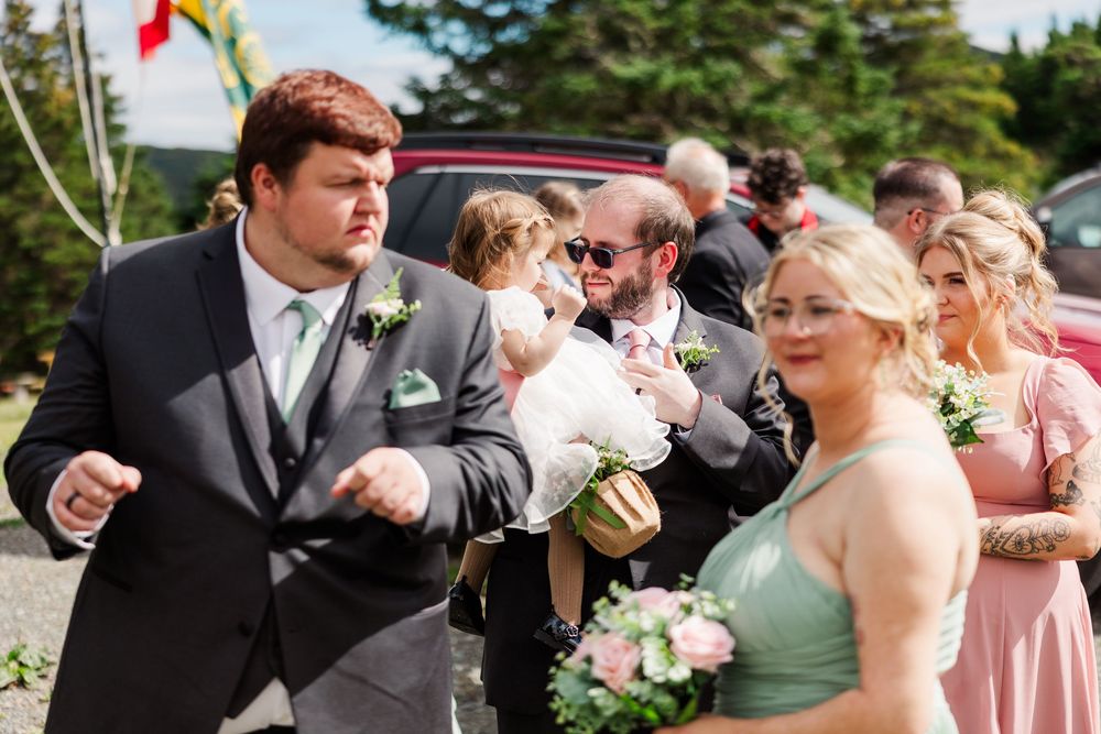 Wedding party members in gray suits and pink bridesmaid dresses gather outdoors, with one man holding a baby in white.