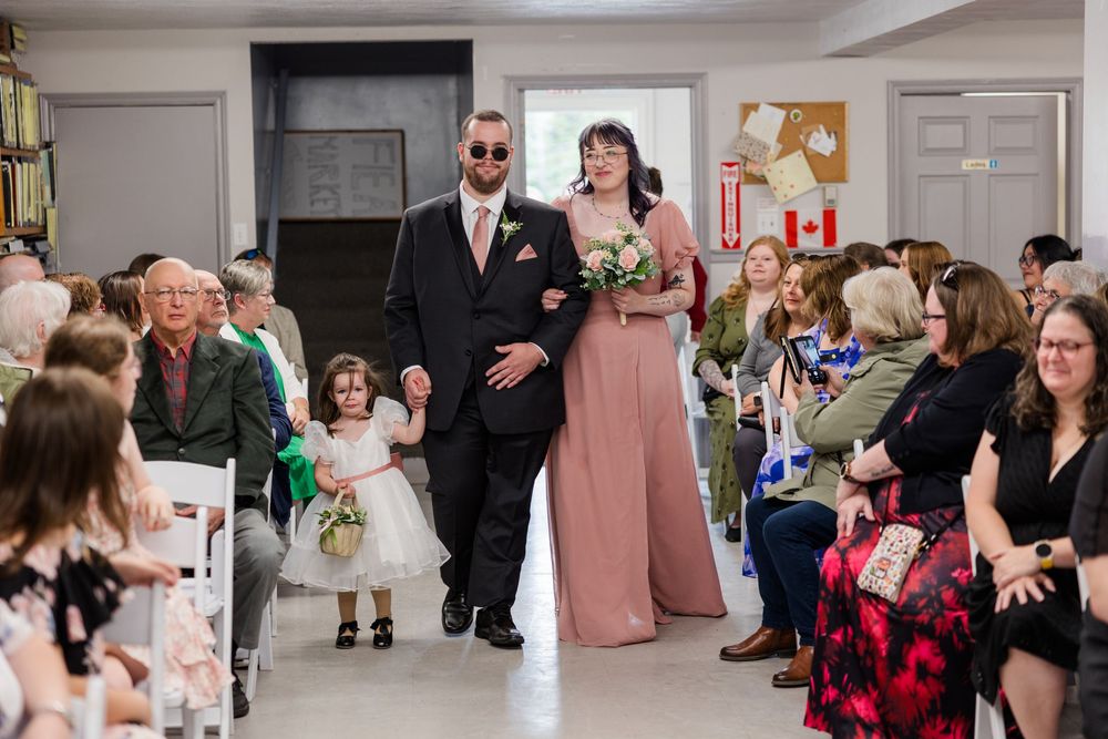 A groomsman in a black suit escorts a bridesmaid in a pink gown down the aisle with a flower girl at a wedding.