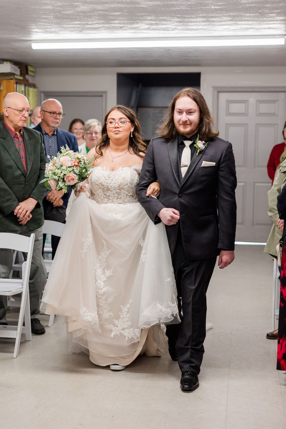 Bride in lace gown and groom in black tuxedo walk down the aisle together during their indoor wedding ceremony.