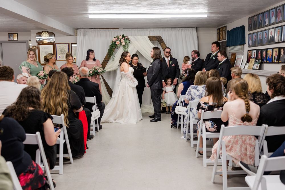Indoor wedding ceremony with bride and groom at the altar, guests seated in white chairs, floral arch backdrop.