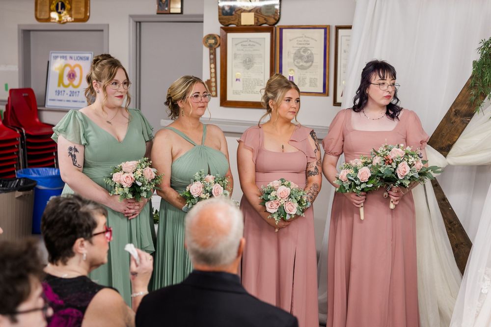 Four bridesmaids in sage green and dusty rose dresses holding pink floral bouquets stand at a wedding ceremony.