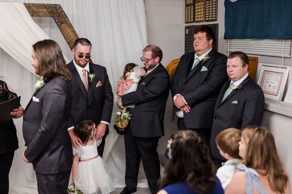 Groomsmen in gray suits with green ties gather indoors during a wedding ceremony, one holding a flower girl.