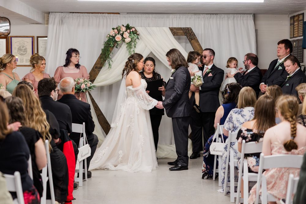 Bride and groom exchange vows at indoor wedding ceremony with geometric arch, bridesmaids, and seated guests.