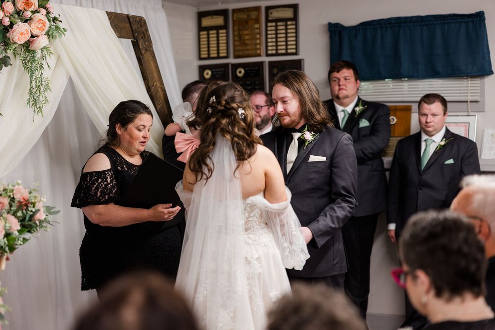 Bride and groom exchange vows at indoor wedding ceremony with floral arch, officiant, and guests in background.