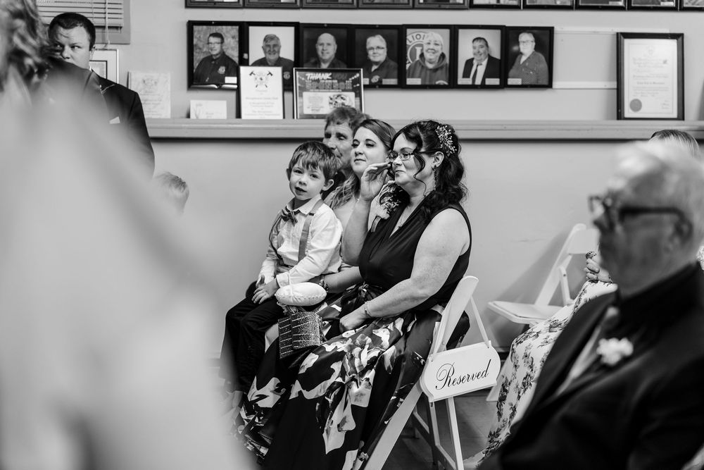 Black and white wedding ceremony photo of guests seated in white chairs, including a woman holding a child.