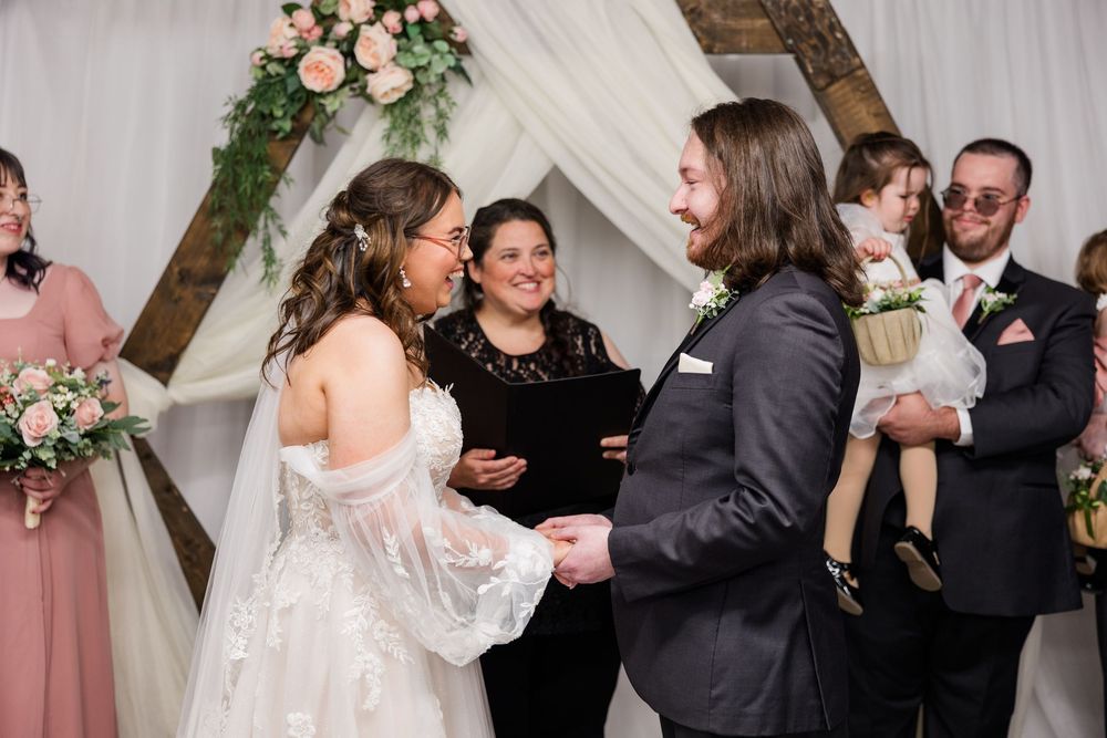 Bride and groom exchange vows at rustic wooden arch altar decorated with pink flowers and white draping.