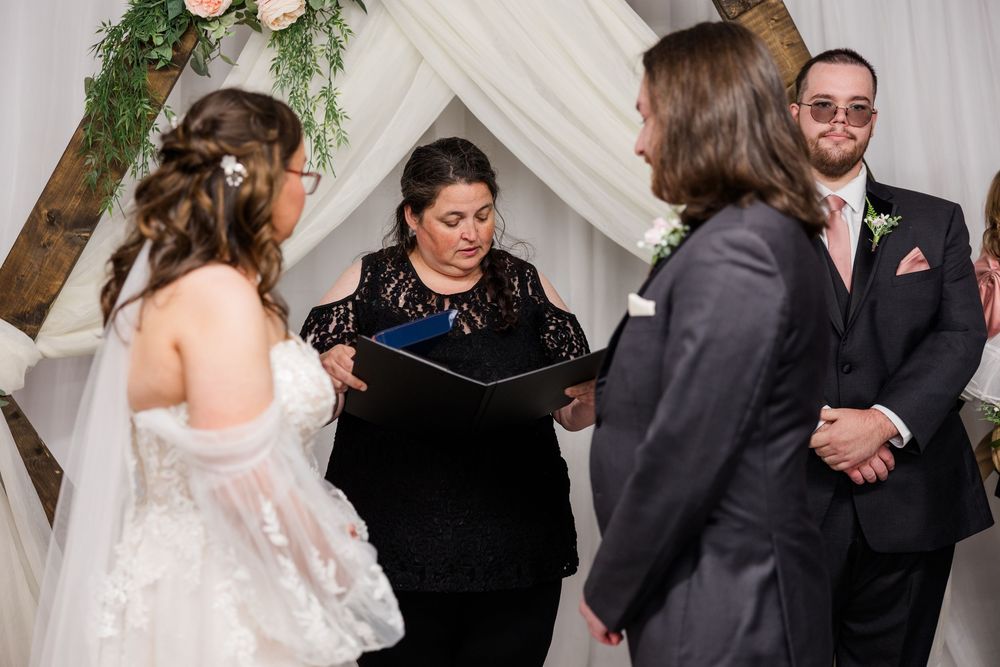 Wedding officiant reads vows as bride and groom stand at a rustic wooden arch altar.
