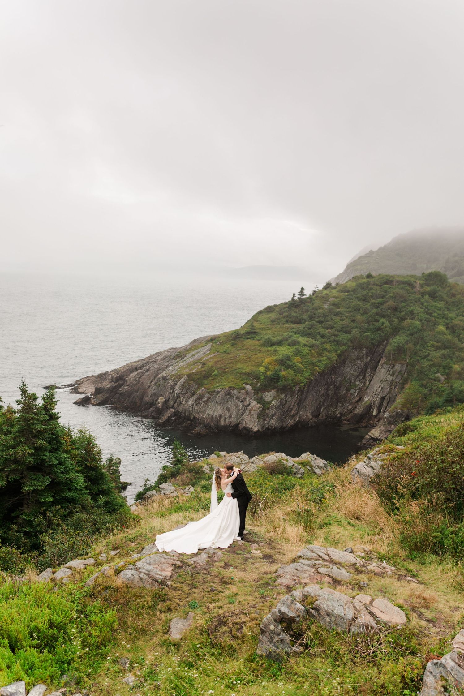 Bride and groom embrace on rocky coastal cliffside with lush greenery and misty ocean backdrop on their wedding day.
