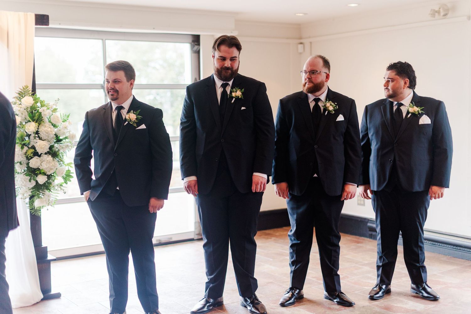 Four groomsmen in dark suits with boutonnieres standing in a bright room near floral arrangements at a wedding ceremony.
