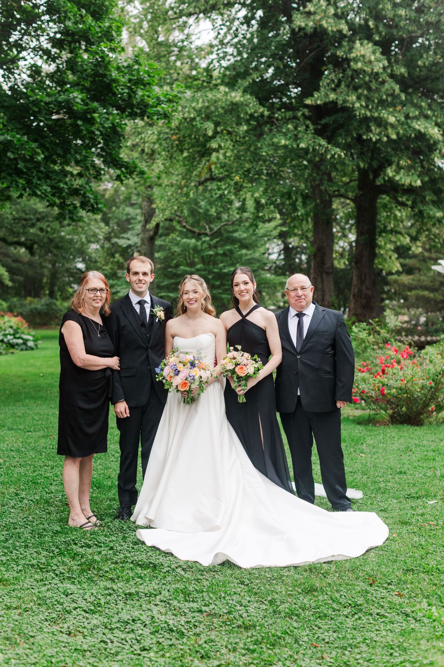 A bride in white gown poses with family in black attire outdoors on green lawn with colorful bouquet.