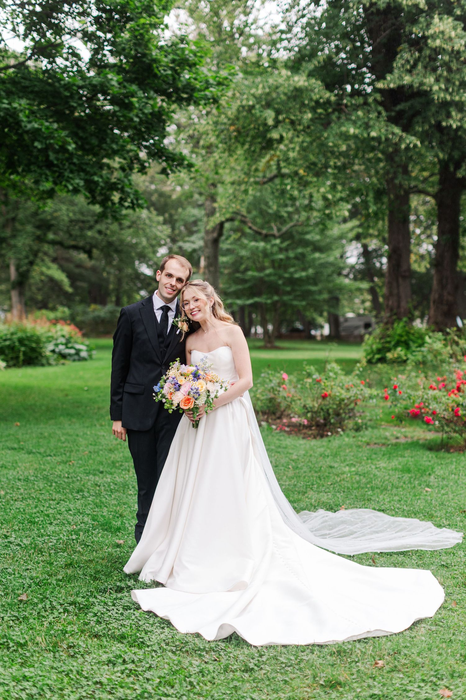 Bride and groom pose together on lush green lawn, bride holding colorful bouquet in elegant white gown with long train.