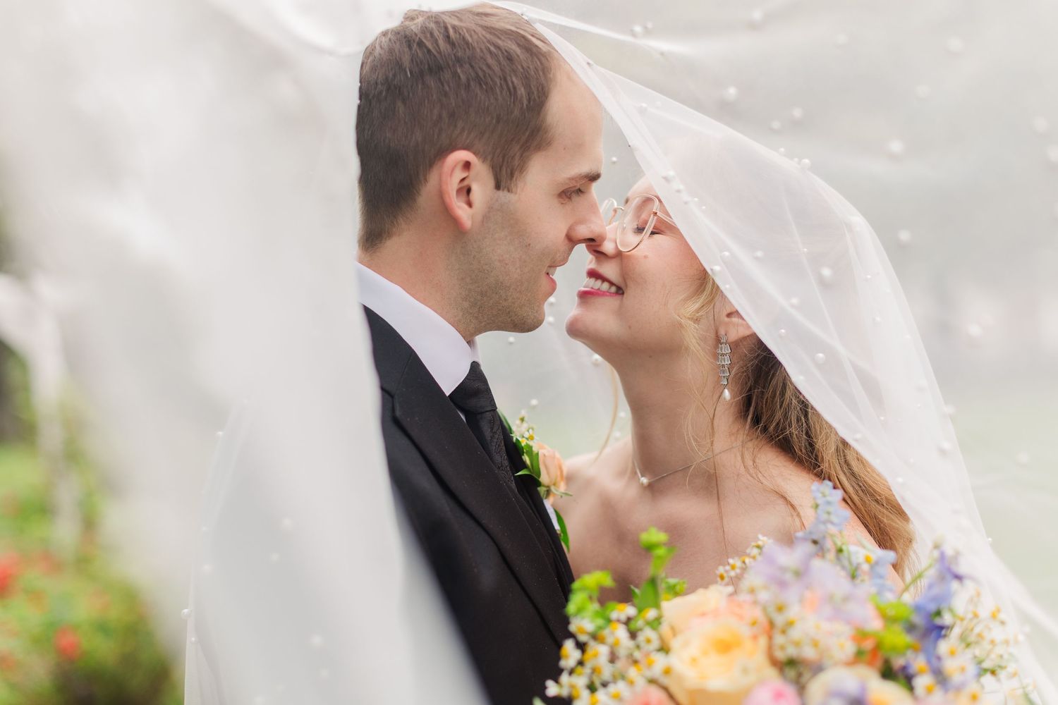 A bride and groom share a romantic moment under a wedding veil, with the bride holding a colorful bouquet.