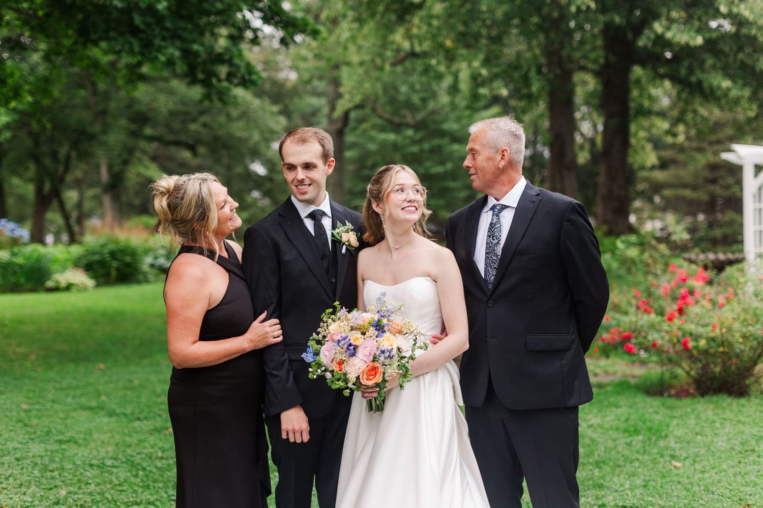 Bride and groom pose with family in lush garden setting, bride holding colorful bouquet in elegant white gown.