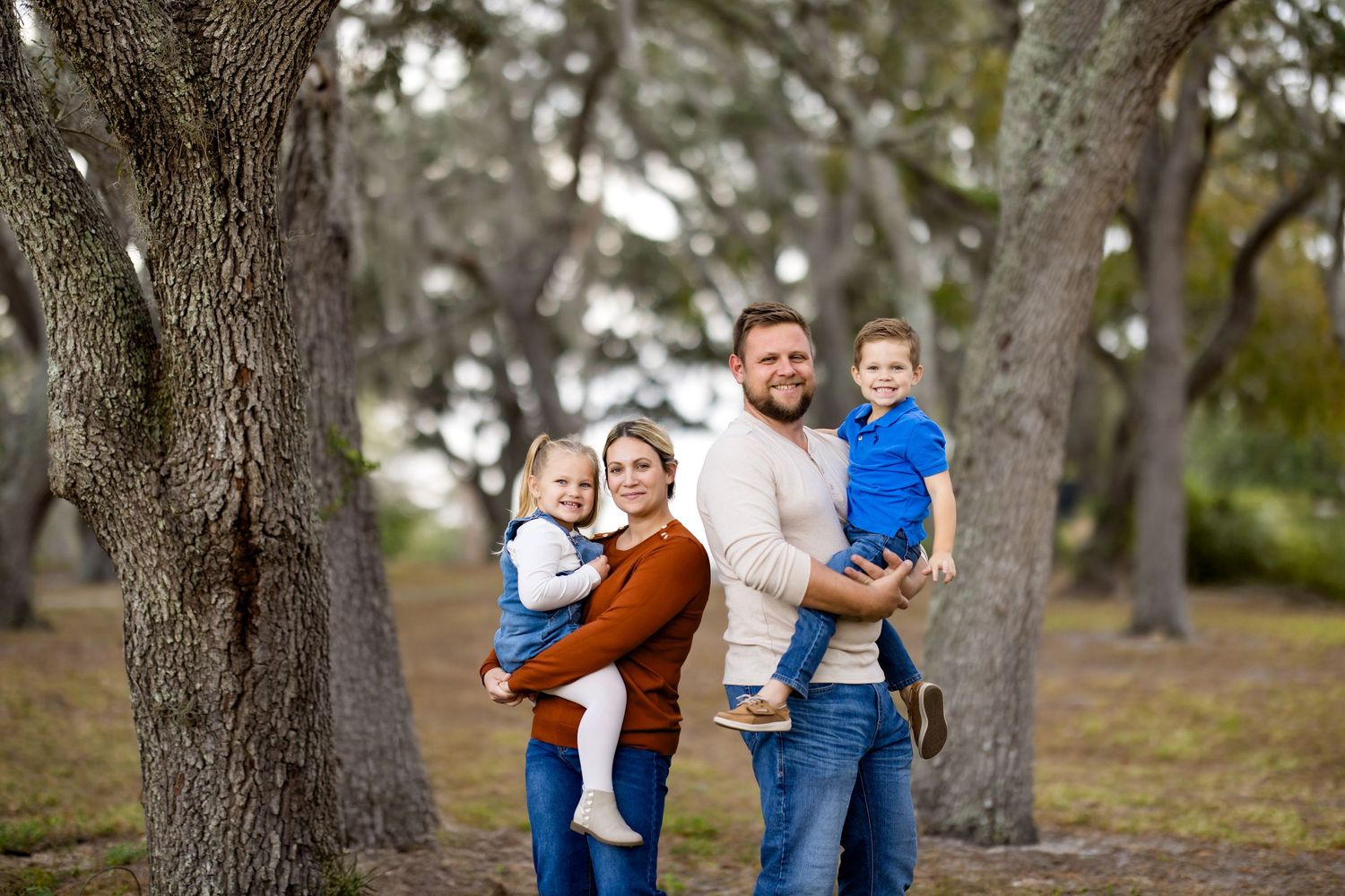 Family of four poses outdoors among moss-covered trees, parents holding young children, smiling for a portrait photo.