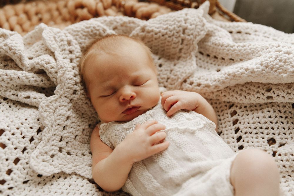 Sleeping newborn in white lace dress lying on cream knit blanket with hands clasped across chest.