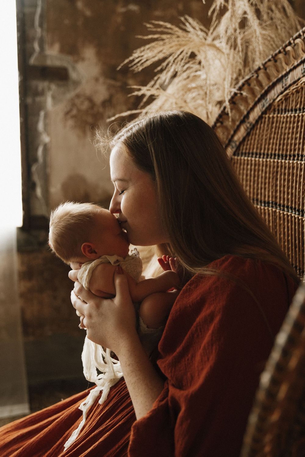 A tender moment captured in warm sepia tones as a mother in a red dress cradles and kisses her infant in a wicker chair.