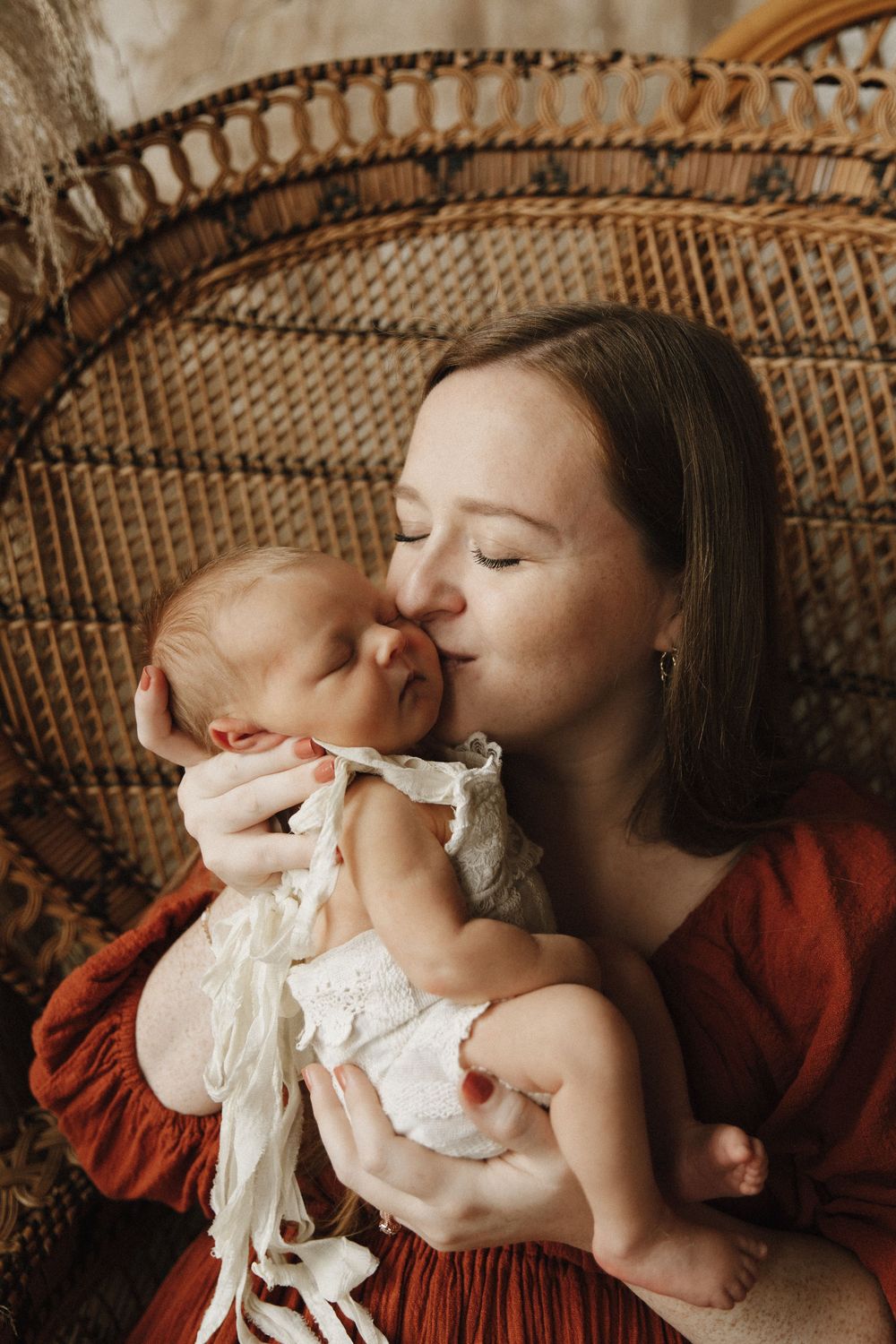 A tender maternal moment captured in warm tones with a vintage wicker peacock chair backdrop.
