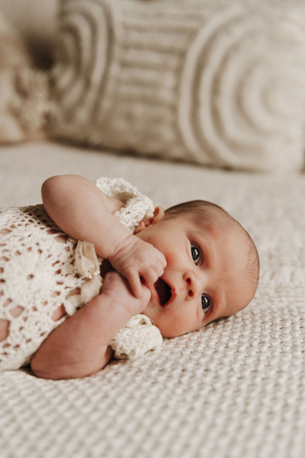 A baby wearing white lace lies on a cream colored knitted blanket in a tender, serene lifestyle photo.