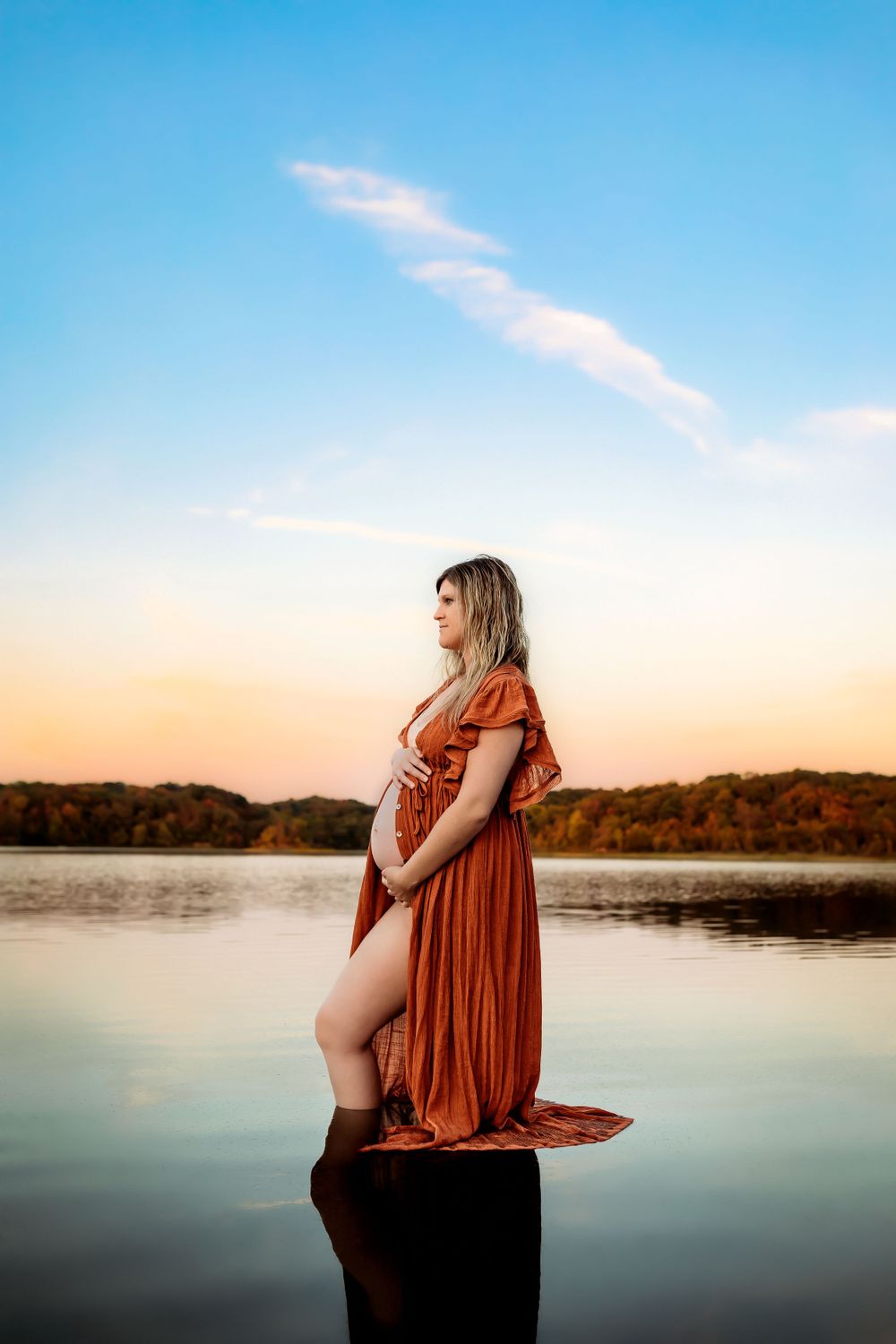 Maternity photo series at sunset shows silhouette against tranquil lake in warm orange and blue tones.
