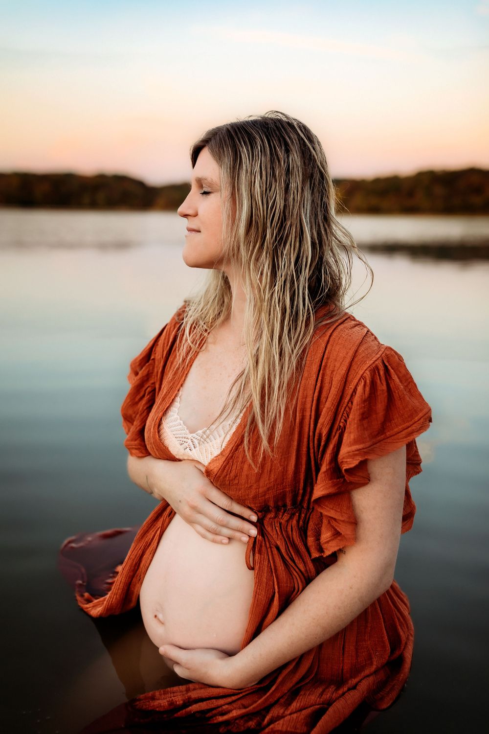 A pregnant woman in a rust-colored dress stands in calm water at sunset, cradling her belly with closed eyes.